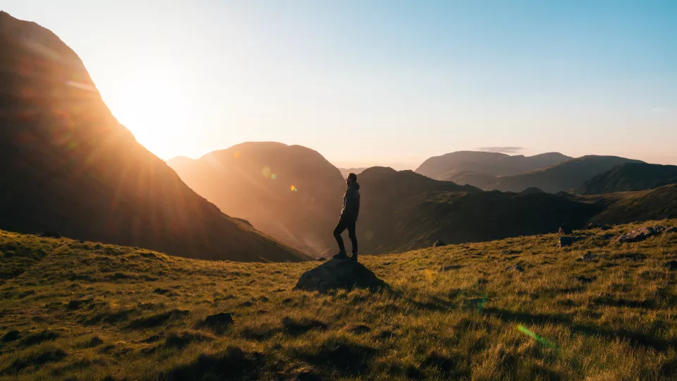 En människa står i ett landskap med berg och solneggång och vänder sig mot solen