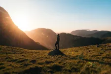 En människa står i ett landskap med berg och solneggång och vänder sig mot solen